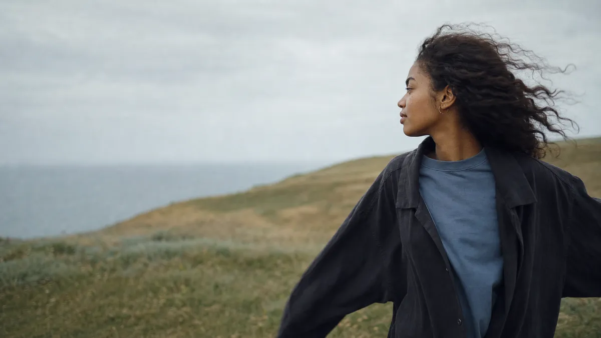 A young woman stands on a windswept coastal hill, hair blowing, looking out over the grey sea under a soft overcast sky.