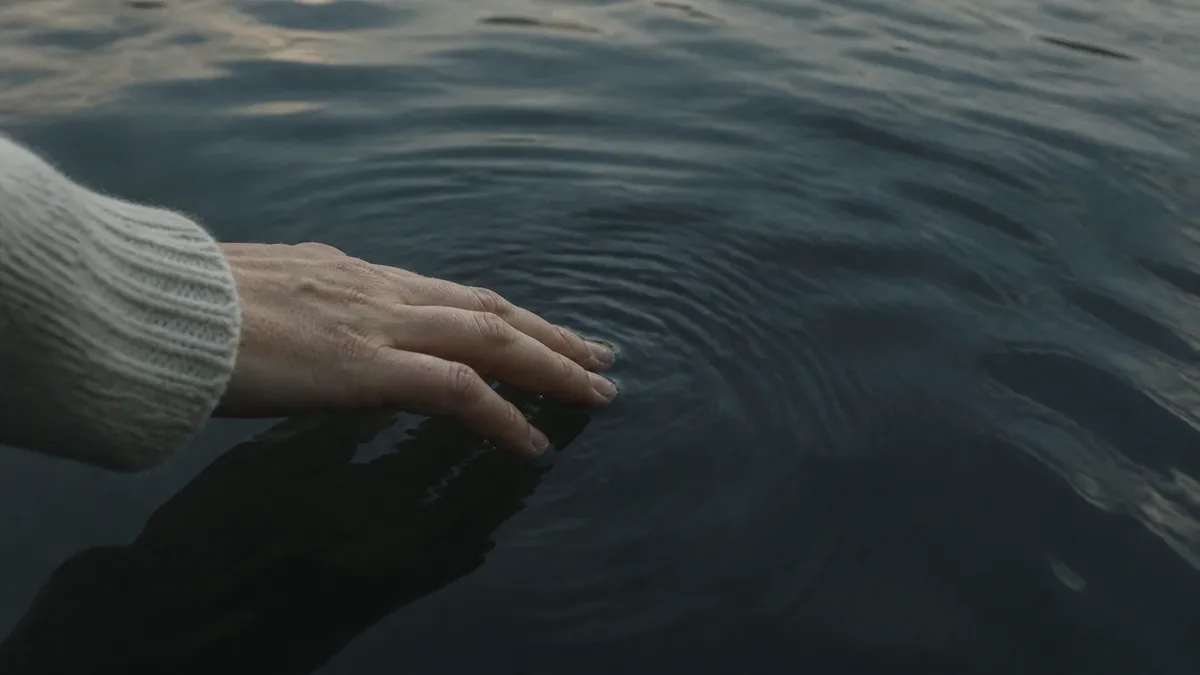 A hand in a cream sweater rests on still dark water, gentle rings rippling outward at dusk.