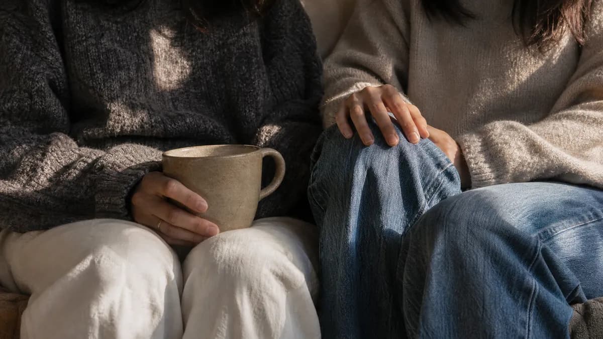 Two people sit close together on a couch in warm light, one cradling a ceramic mug, a hand resting gently on the other’s knee.