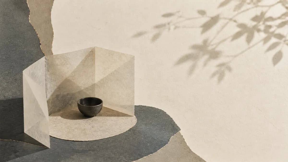 A translucent paper shelter folds protectively around a small dark bowl on a stone plinth, leaf shadows above.