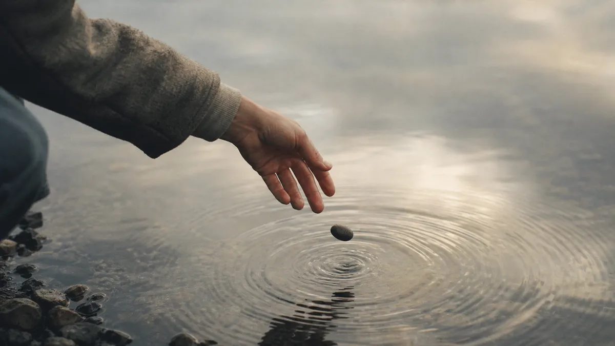 A hand releases a small stone into still water, sending out a single quiet ripple.