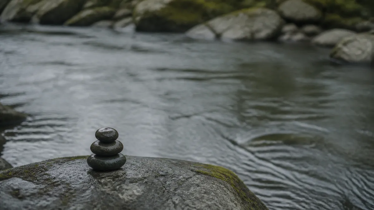 A small cairn of three smooth stones balances on a boulder above a quiet flowing river lined with mossy rocks.