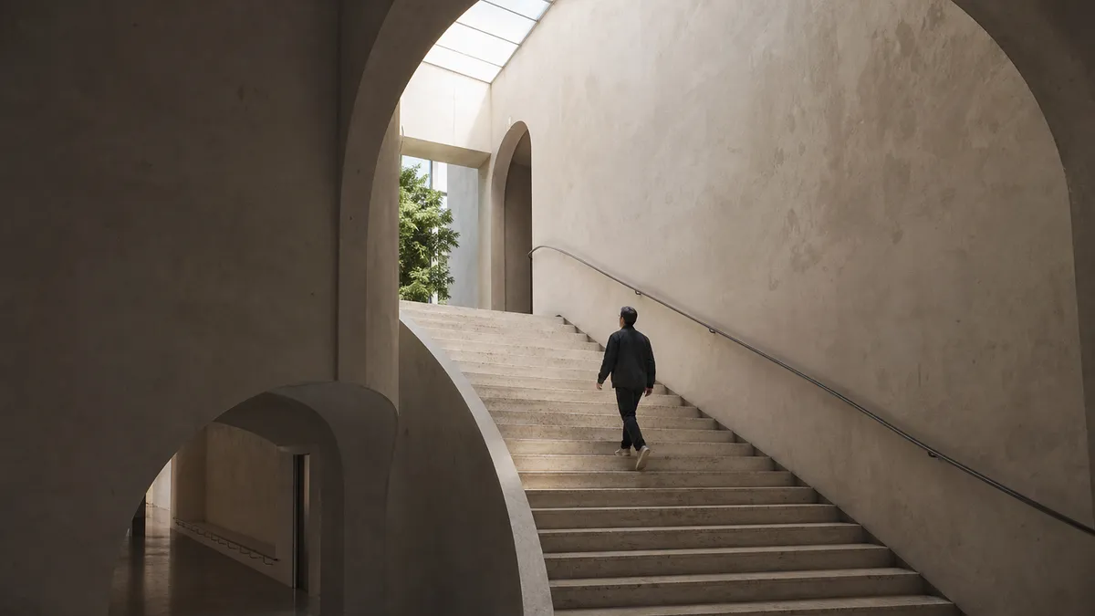 A man climbs a wide stone staircase toward a bright skylight in a vaulted plaster hall, a green courtyard visible through an archway.