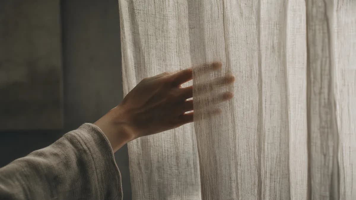 A hand draws back a sheer linen curtain, letting soft daylight into a dim room.