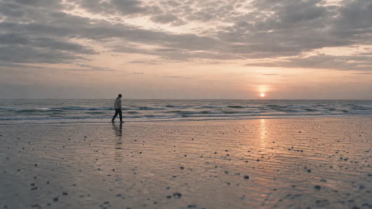 A man walks the wet sand of a wide beach at sunset, small waves rolling in under a pastel sky.