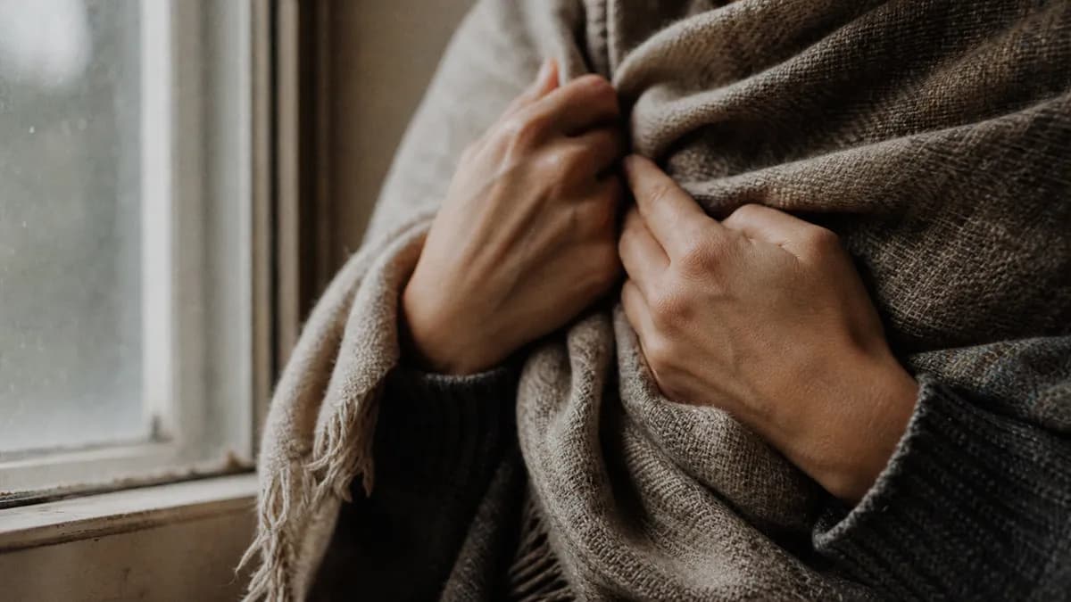 Close-up of two hands drawing a heavy woven blanket tightly around the shoulders beside a frosted window.