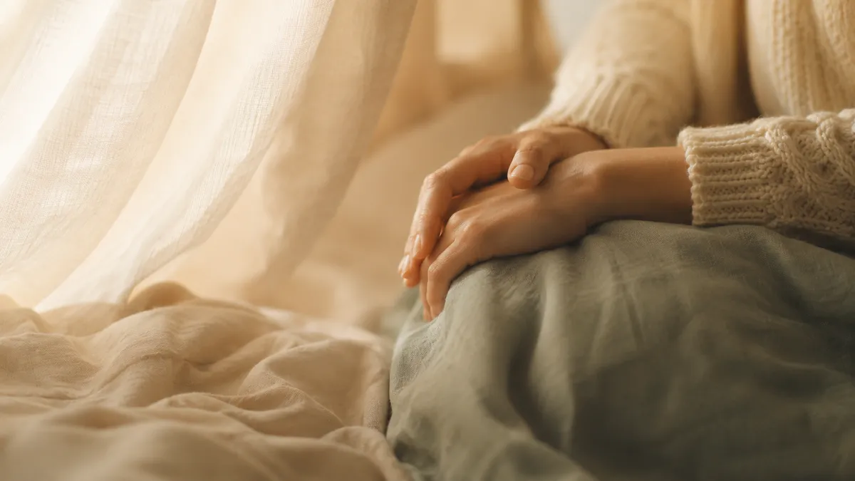A person sits by a window with their hands resting quietly in their lap.