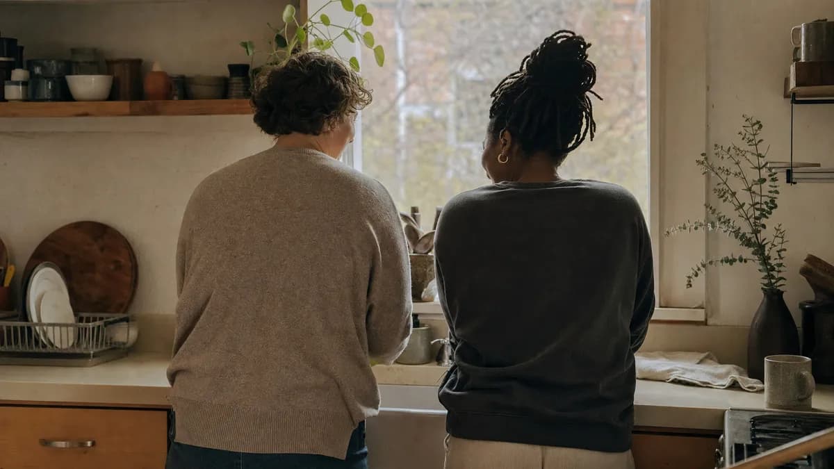 Two people stand side by side at a sunlit kitchen window, quietly busy at the counter together.